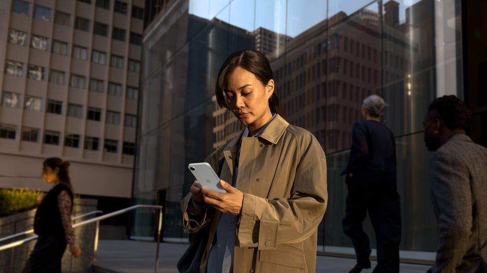 A person using a white iPhone 17e to continue work outside of an office building