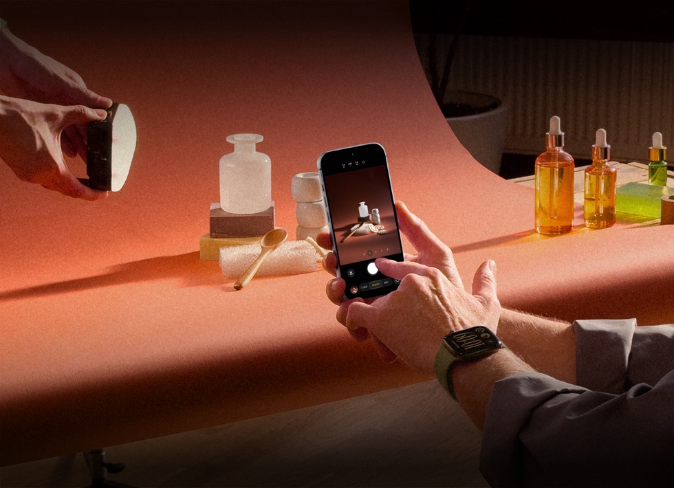 A product photo set, a pair of hands, wearing an Apple Watch device on the left wrist, holds an iPhone 18 Pro, capturing photos of the products, another pair of hands shines a light on the product