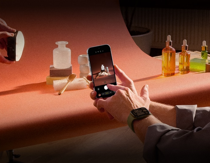 A product photo set, a pair of hands, wearing an Apple Watch device on the left wrist, holds an iPhone 18 Pro, capturing photos of the products, another pair of hands shines a light on the product