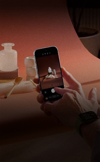 A product photo set, a pair of hands, wearing an Apple Watch device on the left wrist, holds an iPhone 18 Pro, capturing photos of the products, another pair of hands shines a light on the product